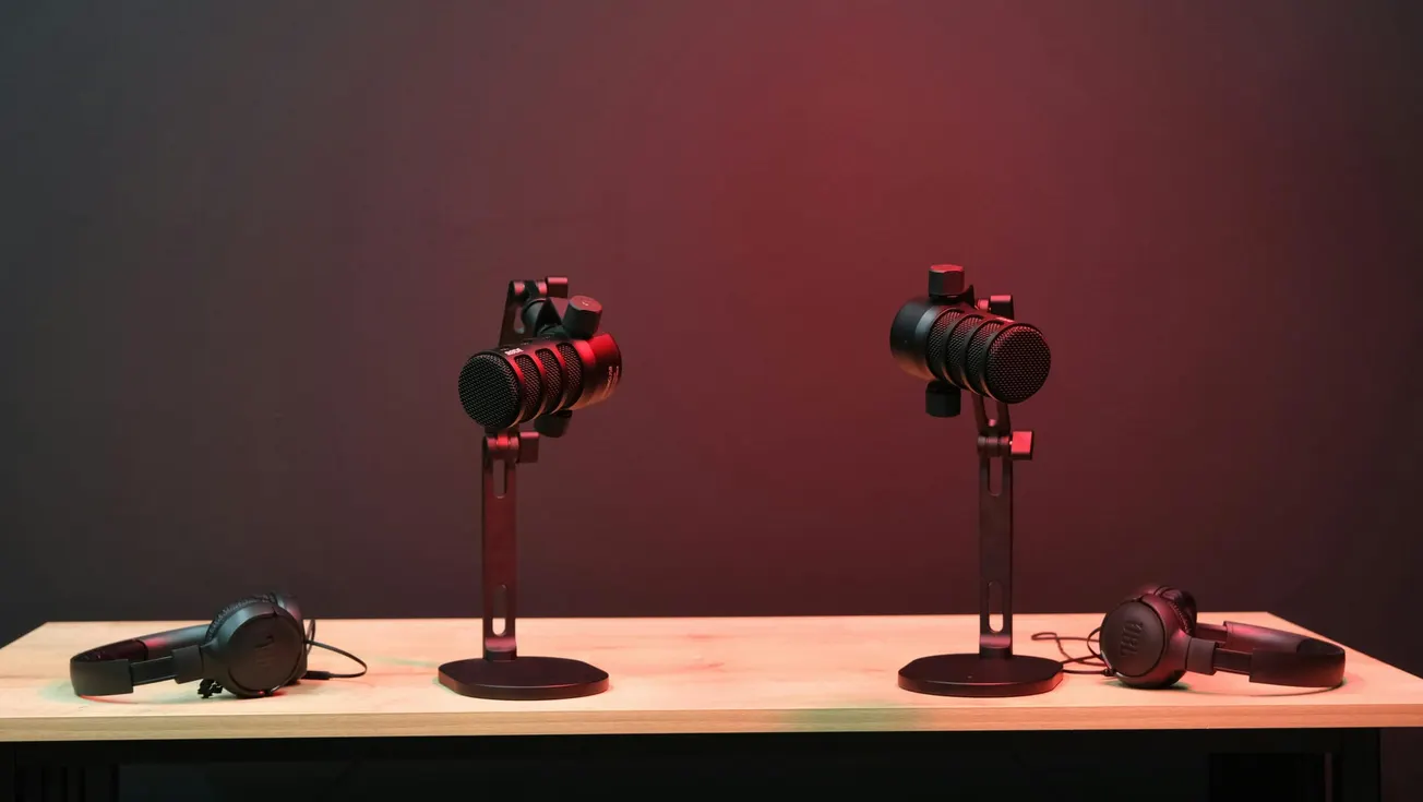 Two microphones on stands are centered on a table under warm red lighting, flanked by a pair of headphones on each side. The scene conveys a professional audio setup.