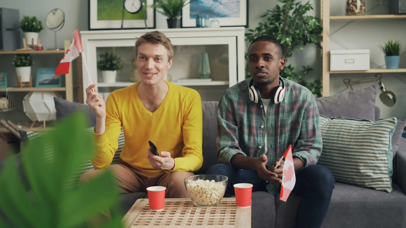 Two men sit on a couch, watching TV with excitement ad holding small Canadian flags. One has headphones around his neck. Popcorn and cups on the table.