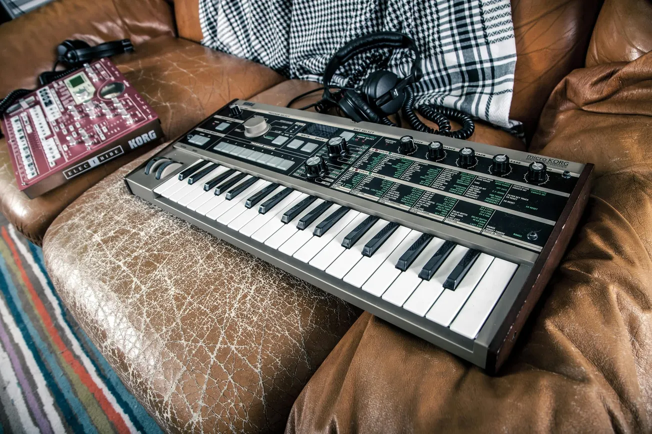 A small synthesizer keyboard with dials rests on a worn brown leather sofa, alongside headphones and another electronic music device. Cozy and creative ambiance.