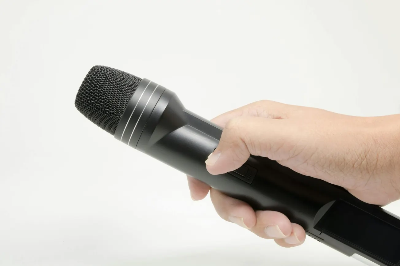A hand holding a black wireless microphone against a plain white background, suggesting an environment ready for speech or performance.