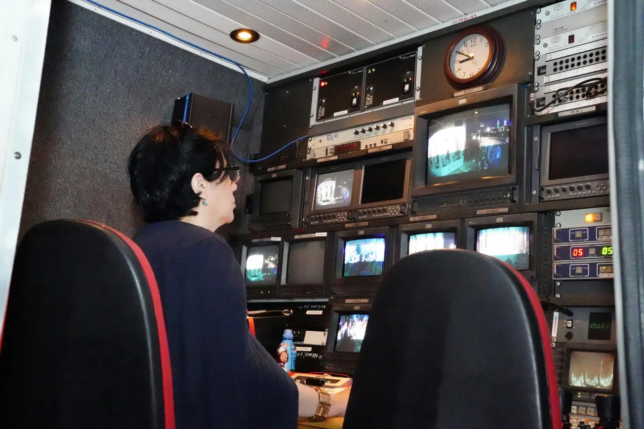 A person sits in a dimly lit broadcast control room, monitoring multiple screens. The equipment-filled environment conveys a focused, technical atmosphere.