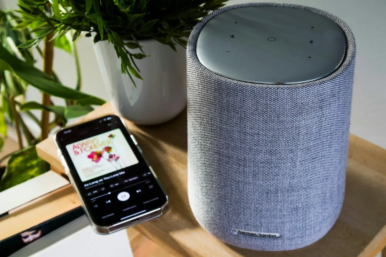 A sleek grey smart speaker on a wooden surface next to a smartphone displaying music. A potted plant adds a touch of greenery in the background.