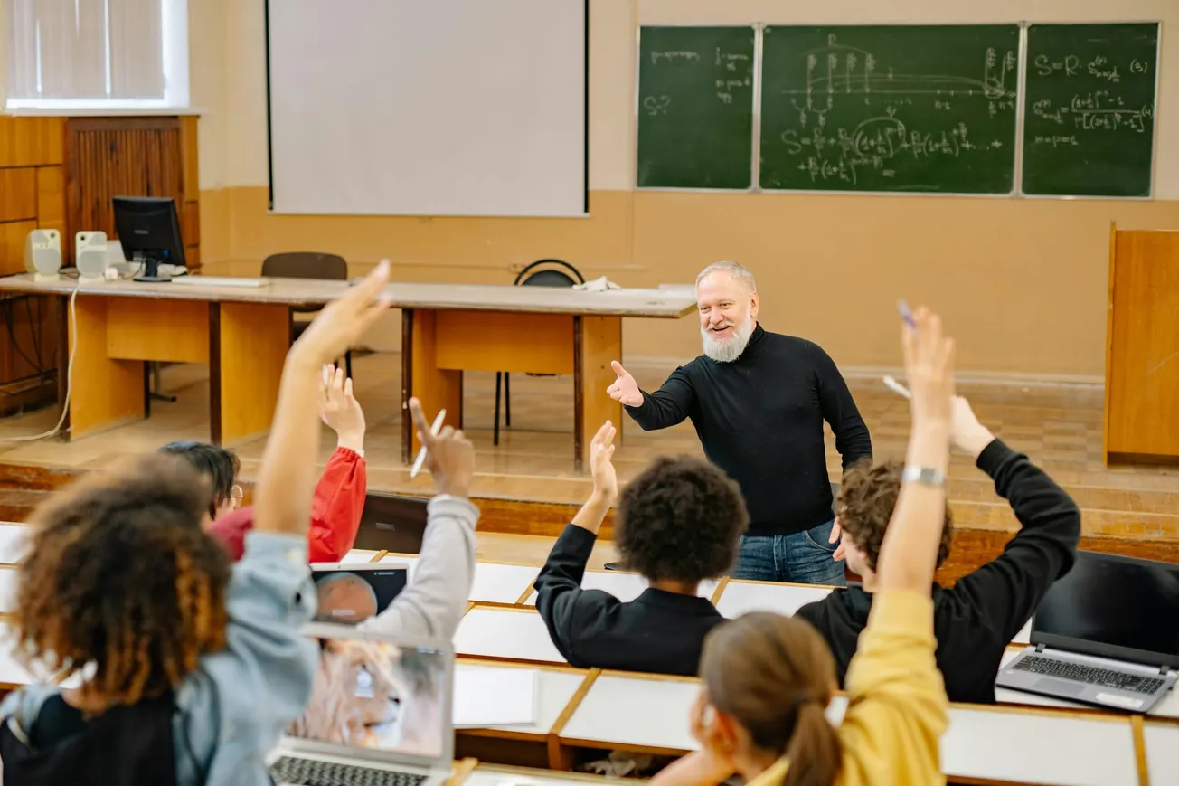 A bearded professor in a black sweater engages with eager students raising hands in a classroom. Chalkboards display complex equations, creating an energetic, academic atmosphere.