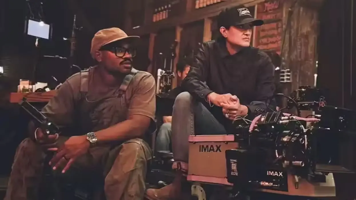 Two filmmakers sit on set beside an IMAX camera. The scene is dimly lit, conveying focus and concentration. One holds a remote, eyes on the scene.