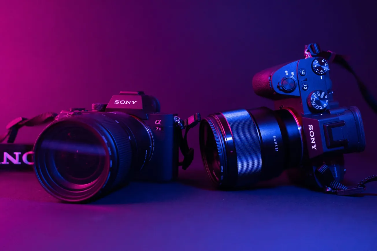 Two Sony cameras with lenses sit on a surface, bathed in moody purple and pink lighting, symbolizing technology and creativity.