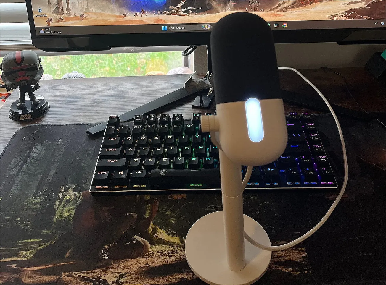 A white and black microphone with a glowing white light sits on a stand in front of a backlit gaming keyboard. A monitor displays a video game.