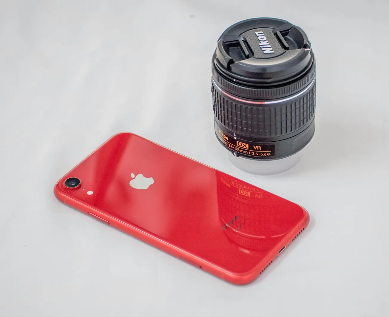 A red smartphone with a white logo lies beside a black Nikon camera lens on a white surface. The scene conveys a tech-savvy and creative vibe.