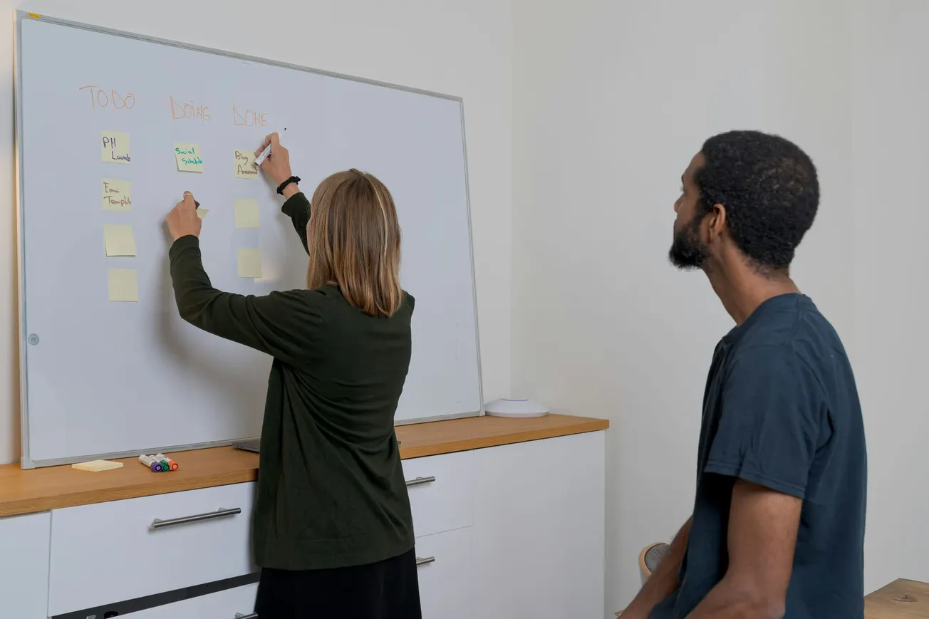 A woman writes on sticky notes on a whiteboard labeled "To Do," "In Progress," and "Done," while a man watches attentively, suggesting collaboration.