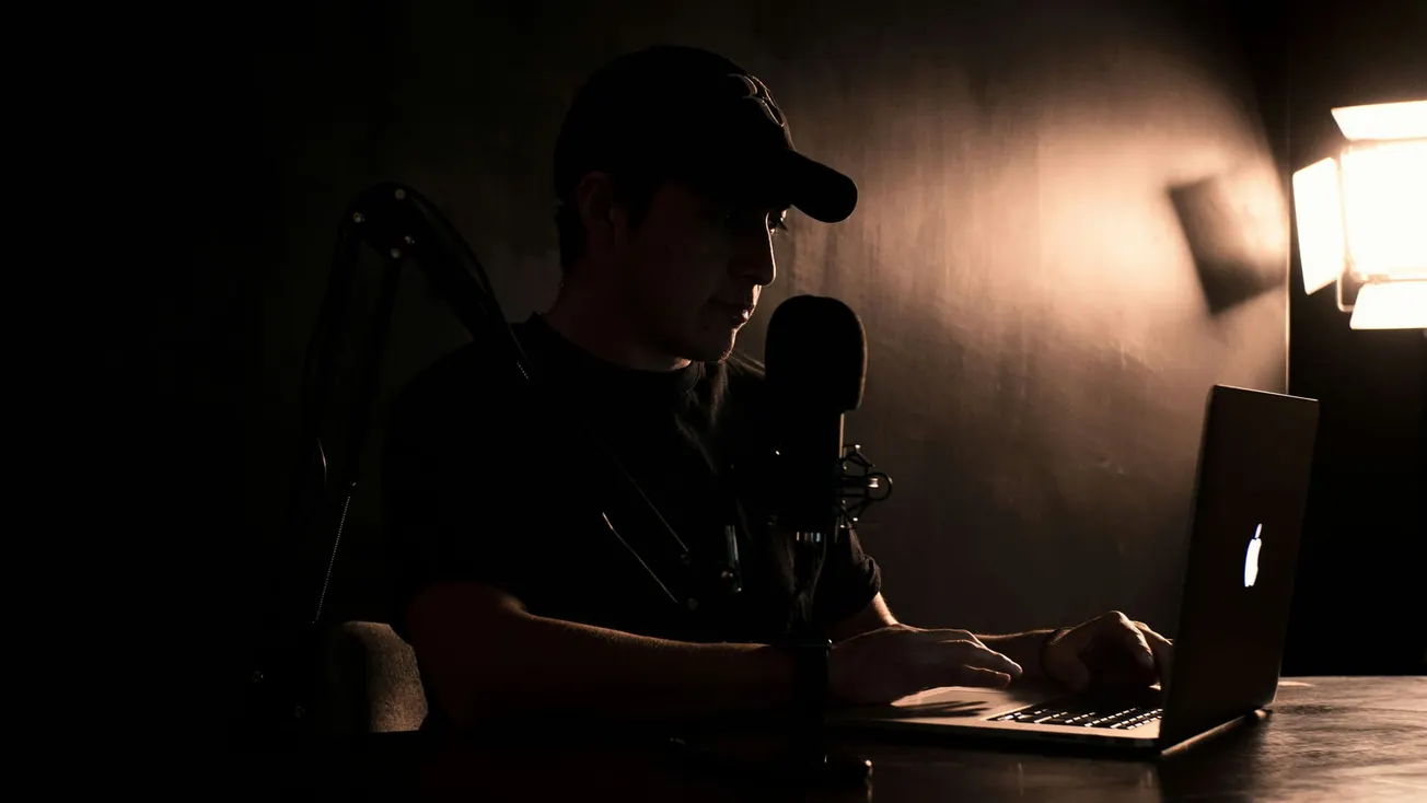 A person in a dark room works at a laptop, lit by a glowing screen and a soft overhead light. A microphone and stand are visible, suggesting podcasting.