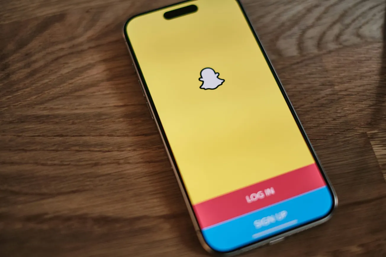 Smartphone on a wooden surface displaying the Snapchat login screen. The background is bright yellow with the Snapchat ghost logo, and options to log in or sign up.