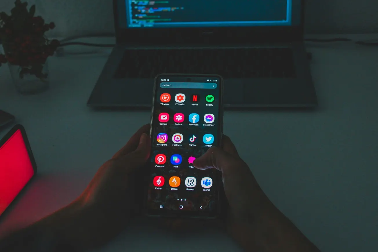 Hands holding a smartphone displaying colorful app icons in a dim room, with a laptop in the background, creating a focused and tech-savvy atmosphere.