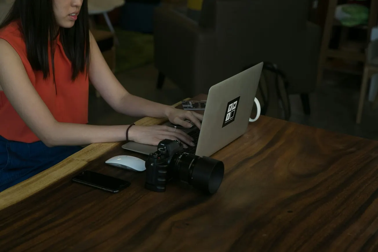 A woman in a red top uses a laptop at a wooden table, alongside a camera, smartphone, and mouse in a cozy, modern setting, conveying focus and creativity.