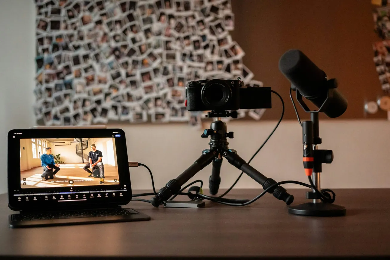 Laptop, camera on a tripod, and microphone on a desk setup for content creation. Blurred background of a large polaroid photo collage adds a creative touch.