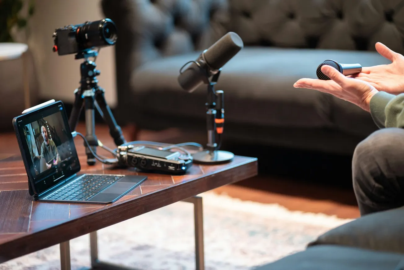 A podcast recording setup with a laptop showing a video call on a wooden table. Nearby are a microphone, audio interface, and camera on a tripod.