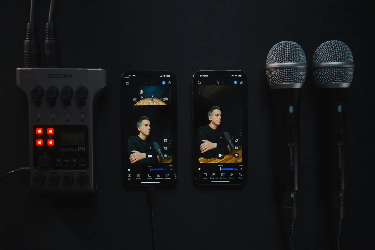 Audio equipment setup with two smartphones displaying a man speaking into a microphone. Nearby are two microphones and a Zoom PodTrak P4 recorder.