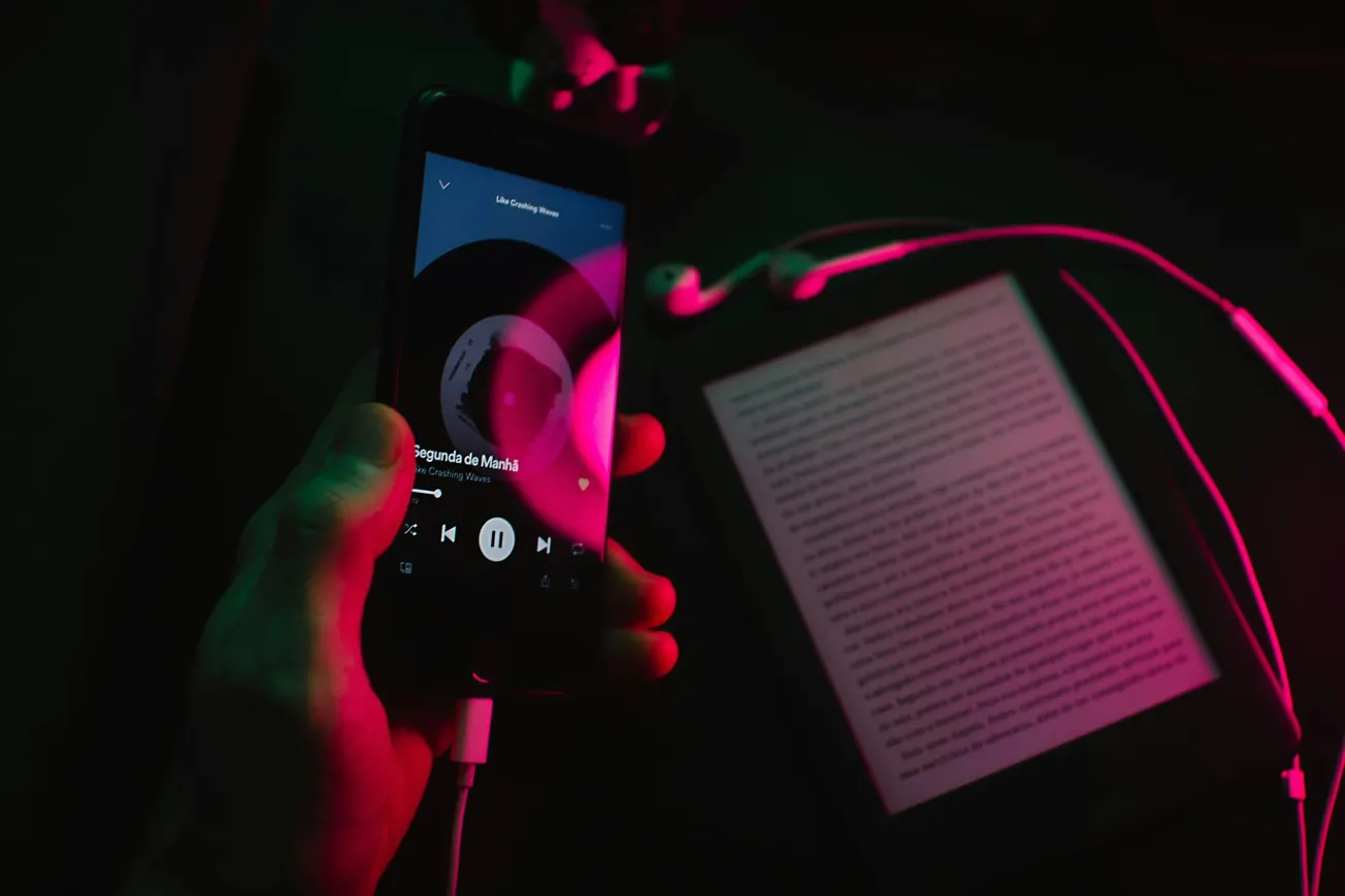A person holds a smartphone displaying a music app, glowing in pink and green light. Nearby are earphones and an e-reader on a dark surface.