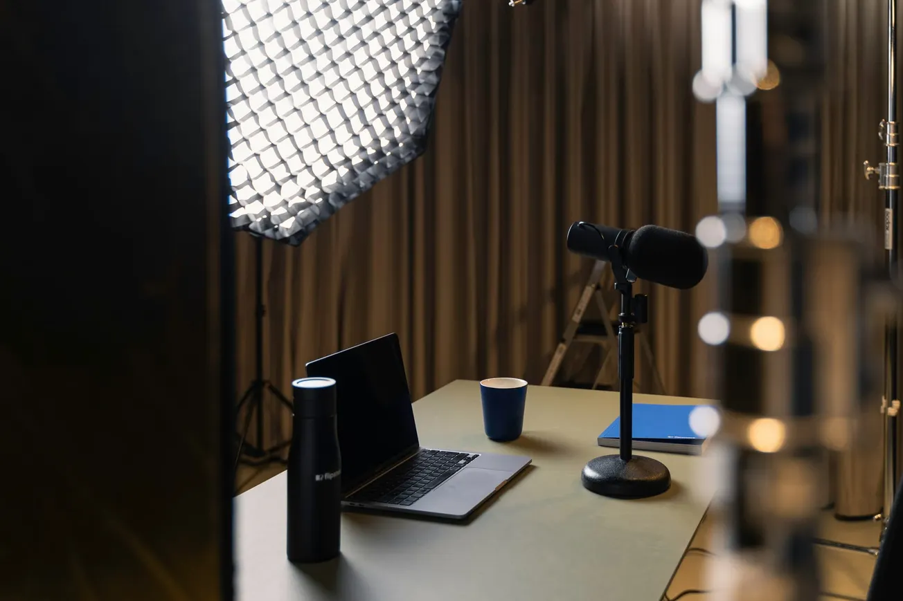 A podcast studio setup with a microphone on a stand, laptop, cup, and bottle on a desk. Soft lighting and a curtain backdrop create a cozy atmosphere.