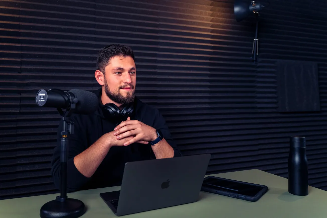 A man sits in a soundproof booth with a microphone, laptop, headphones, and water bottle. He appears thoughtful, ready to record or stream.