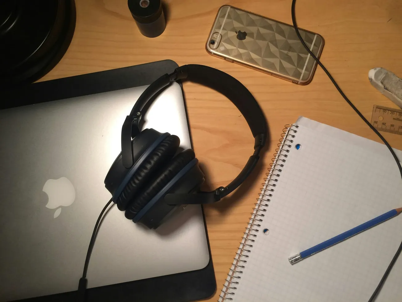 A wooden desk with a closed laptop and black headphones on top. Nearby, there's a patterned smartphone, a spiral notebook, and a pencil, creating a cozy study vibe.