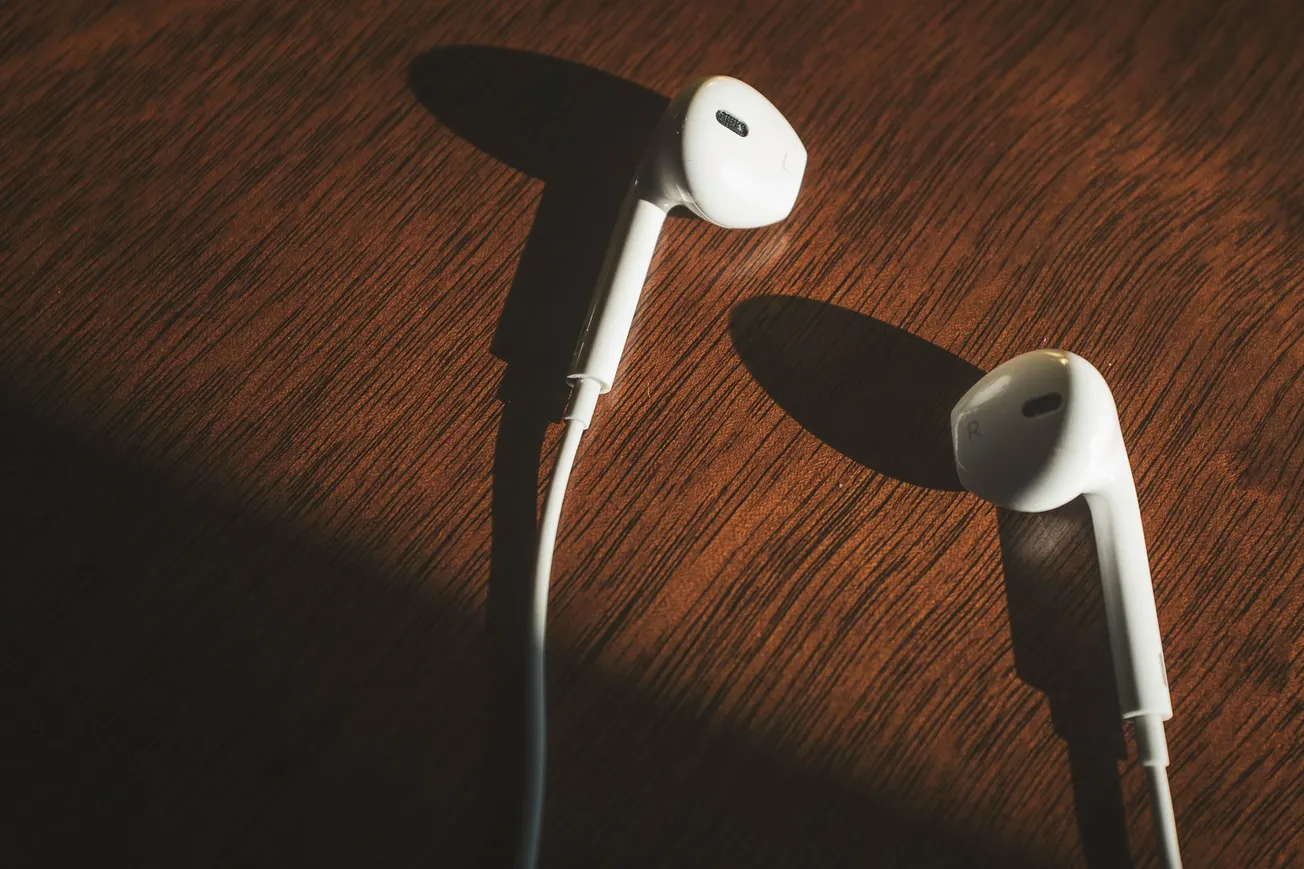 White earbuds lie on a wooden surface, lit by warm sunlight, casting distinct shadows. The scene conveys a calm, minimalist vibe.