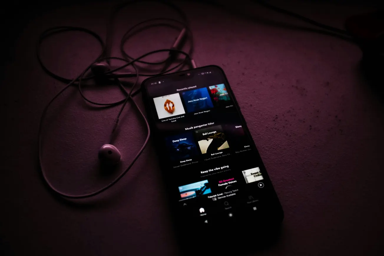 A dimly lit scene showing a smartphone displaying a music app, surrounded by white earphones. The mood is relaxed and intimate, with focus on music exploration.