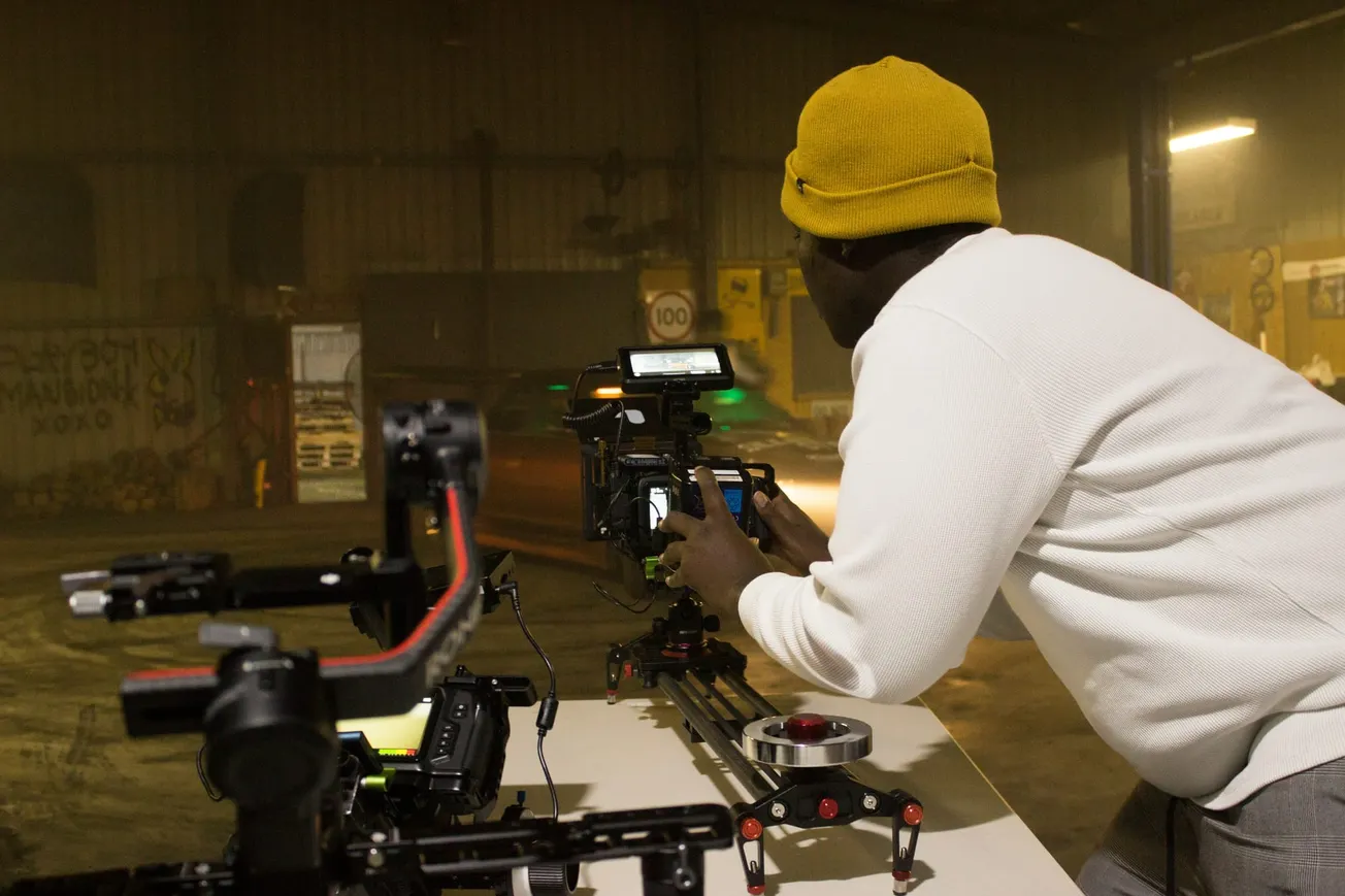 A person in a yellow beanie is operating a camera on a track in a dimly lit warehouse. The scene is focused and professional, with a creative atmosphere.