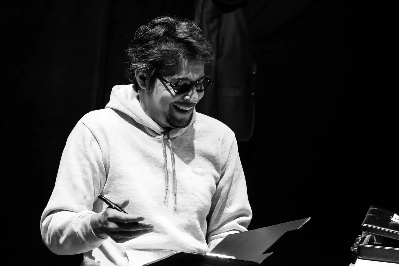 A man in glasses and a hoodie sits at a table, smiling while holding a pen and paper. The black-and-white image conveys a relaxed, joyful mood.