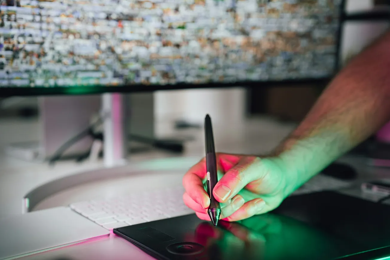 A person uses a stylus on a digital tablet, with a colorful computer screen in the background. The scene is softly lit with green and pink hues.