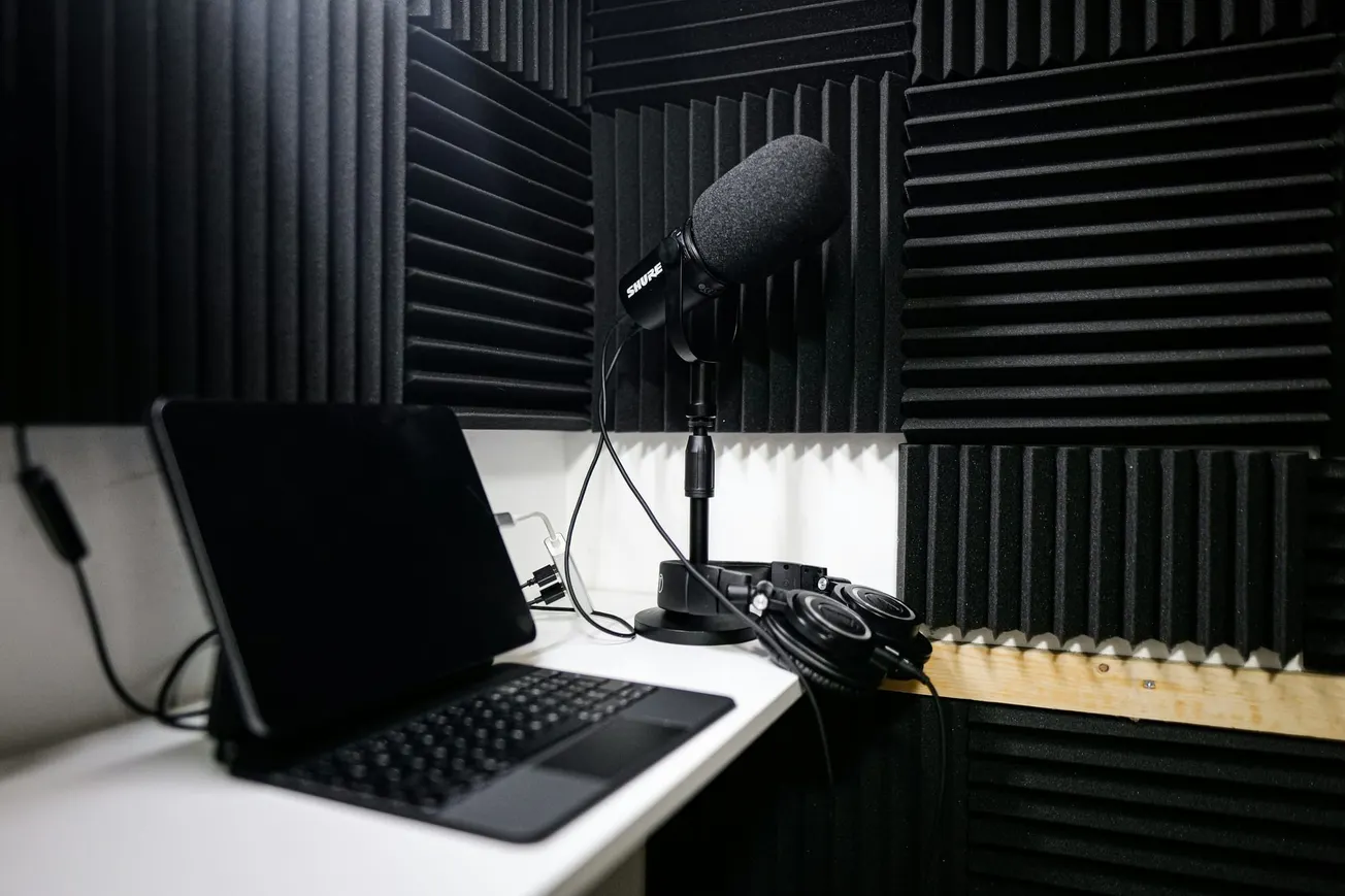 A recording studio with soundproofing foam on walls, featuring a microphone on a stand, a laptop, and headphones on a desk, creating a focused atmosphere.
