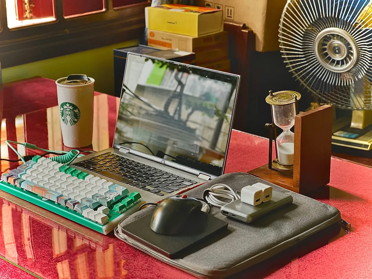 A cozy workspace with a laptop and colorful keyboard. A Starbucks cup, hourglass, and fan are on the desk, creating a warm, relaxing atmosphere.