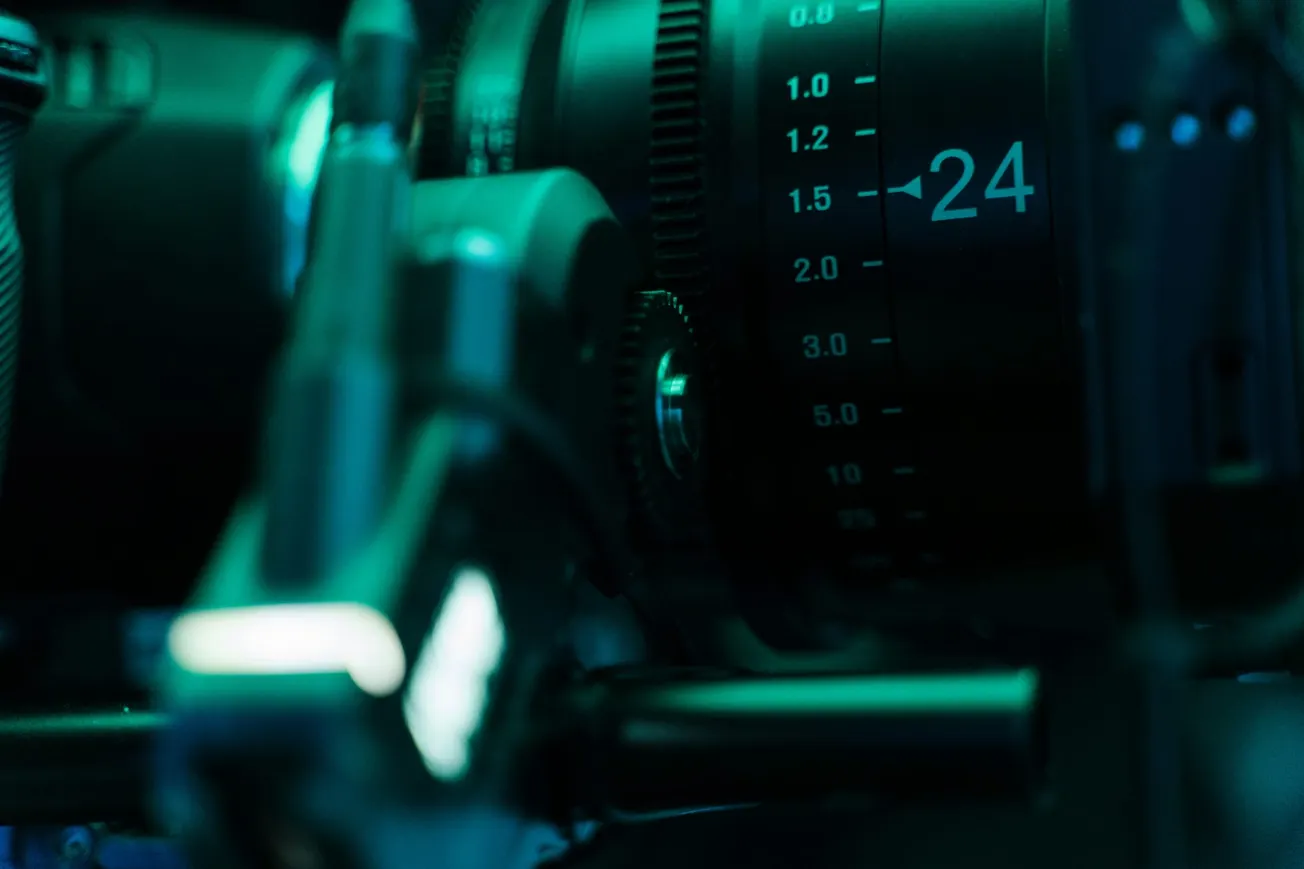 Close-up of a camera lens with focus markings under green lighting, conveying a high-tech, cinematic feel. Numbers and mechanical details are prominent.