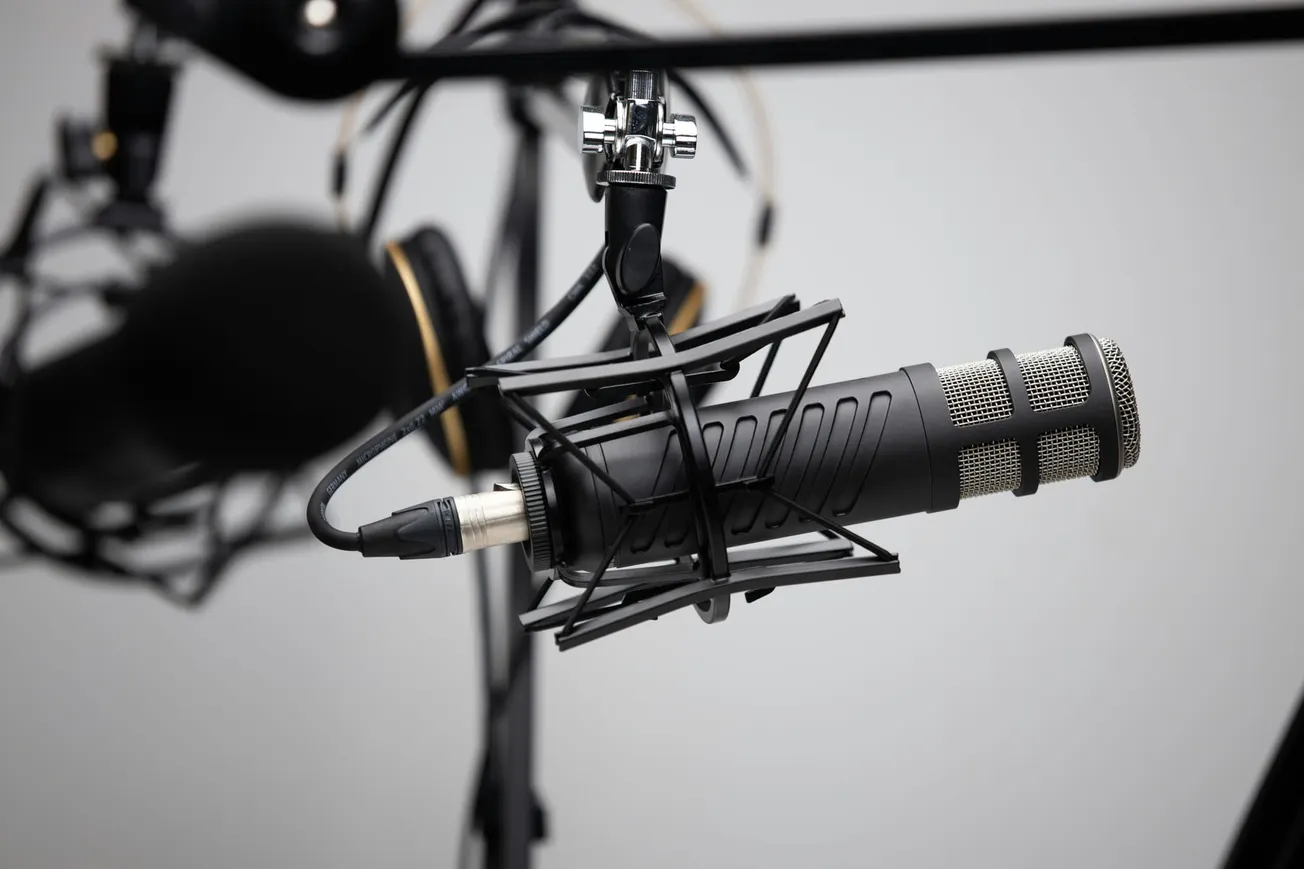 Close-up of a professional microphone in a shock mount, with blurred headphones in the background. The setting feels technical and focused.