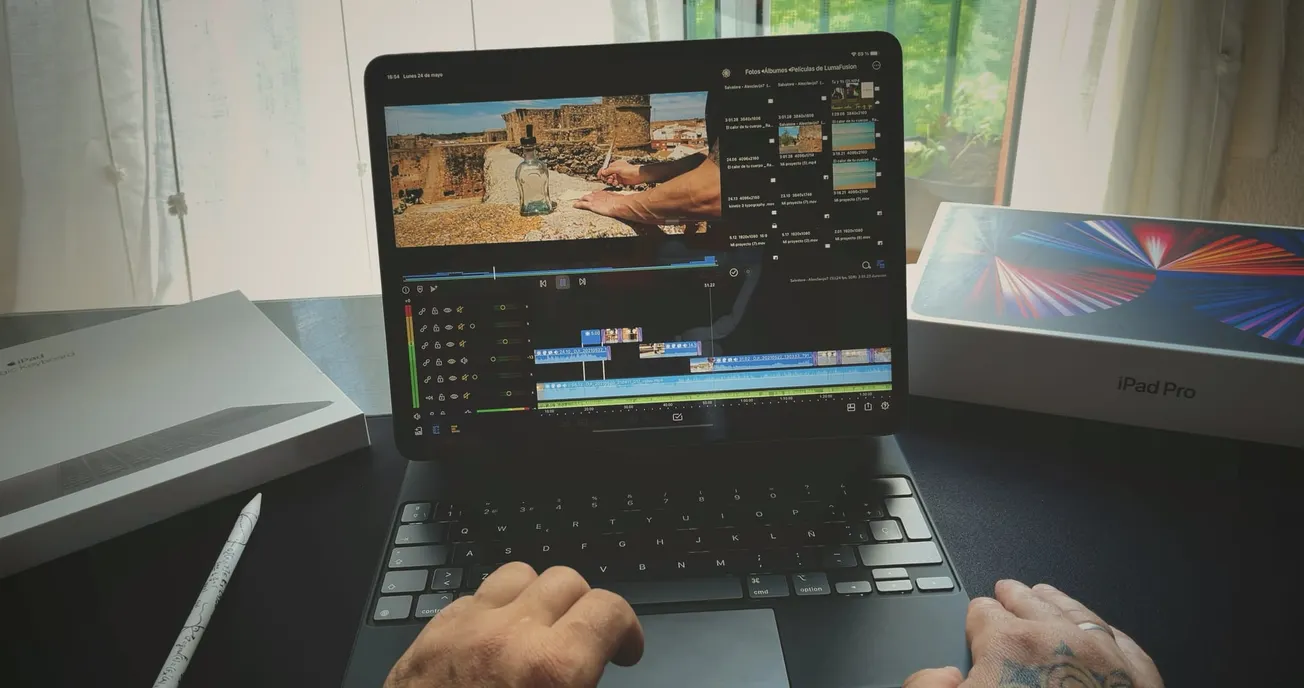Hands editing video on an iPad Pro with keyboard. A stylus and boxes are nearby. Scene through a window gives a relaxed, creative vibe.