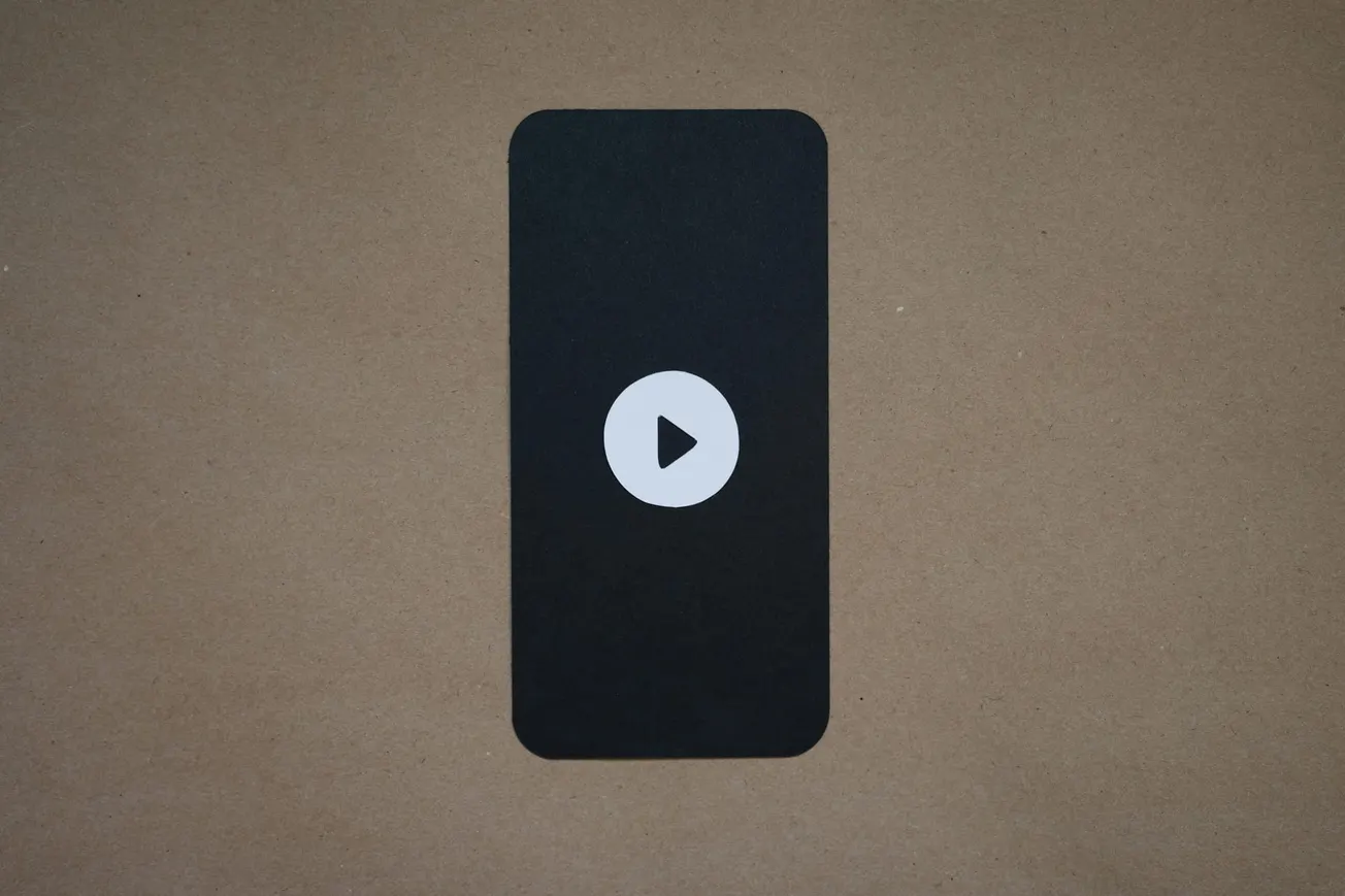 A black rectangle with rounded corners on a brown background features a white play button in the center, resembling a smartphone screen.