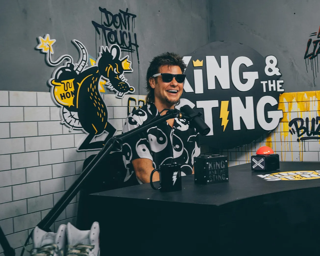 Theo Von in sunglasses sits at a podcast desk with a black and white yin-yang shirt. The background has graffiti-style art and "King & the Sting" text. Playful vibe.