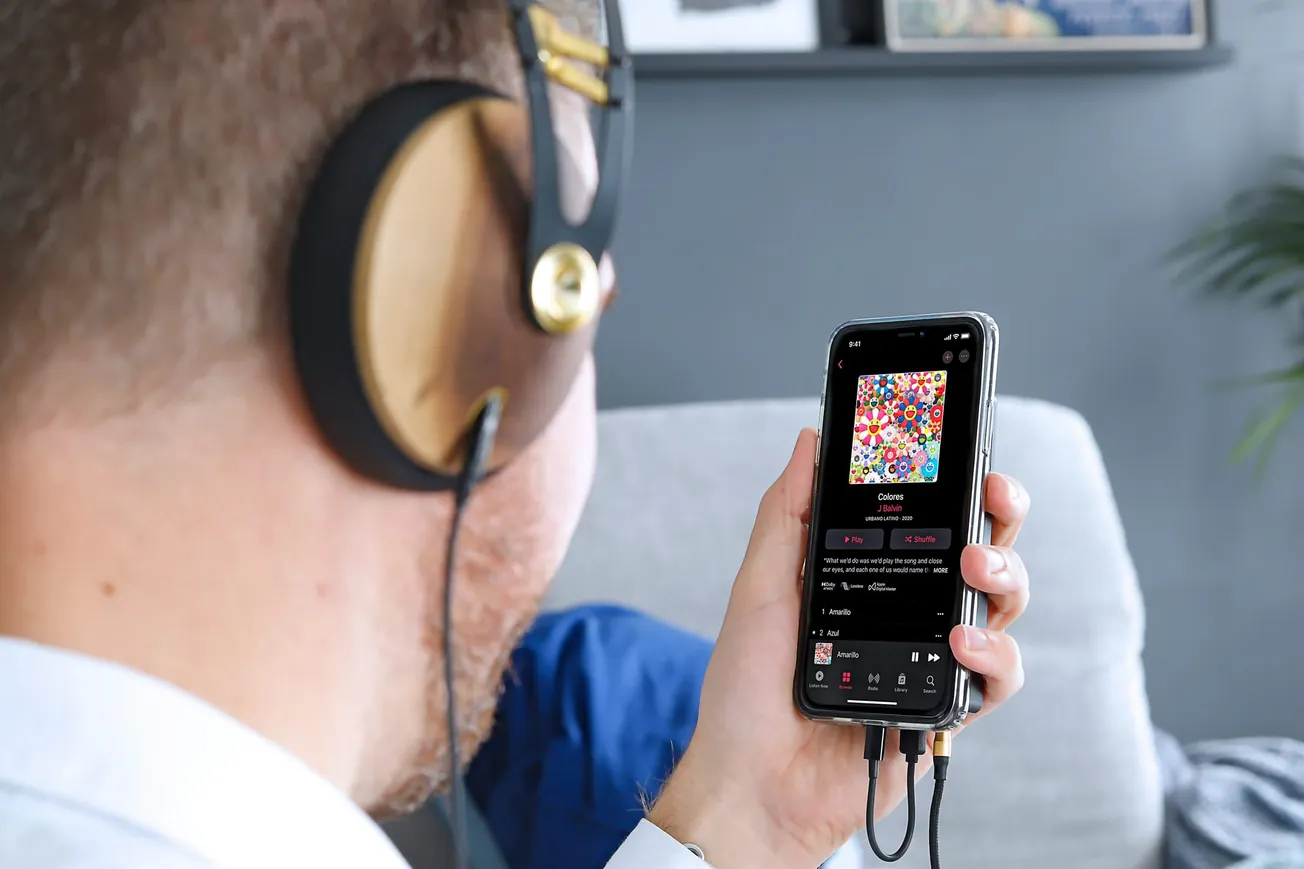 A person wearing large headphones listens to music on a smartphone displaying an album with colorful art. The setting is a casual living room.