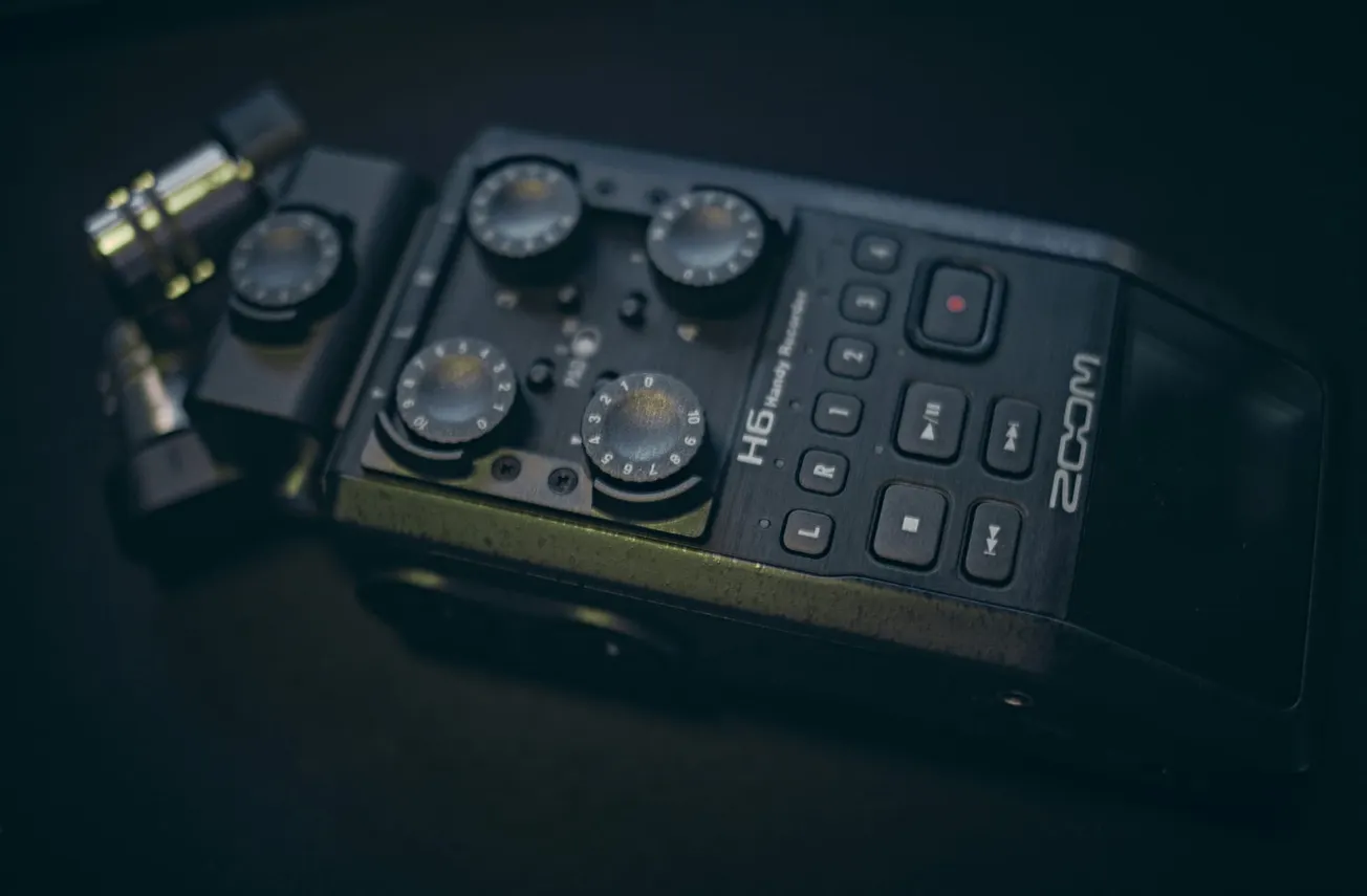 Close-up of a Zoom H6 Handy Recorder on a dark surface. The device features four round control knobs and multiple buttons, conveying a sense of professional audio equipment.