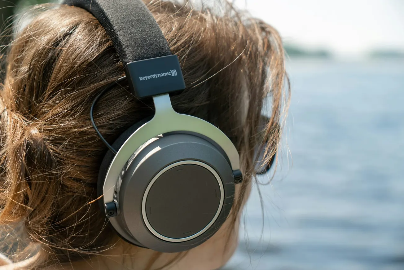 A person with brown hair wears large, over-ear headphones while facing a blurred, serene lakeside view. The scene conveys relaxation and escape.