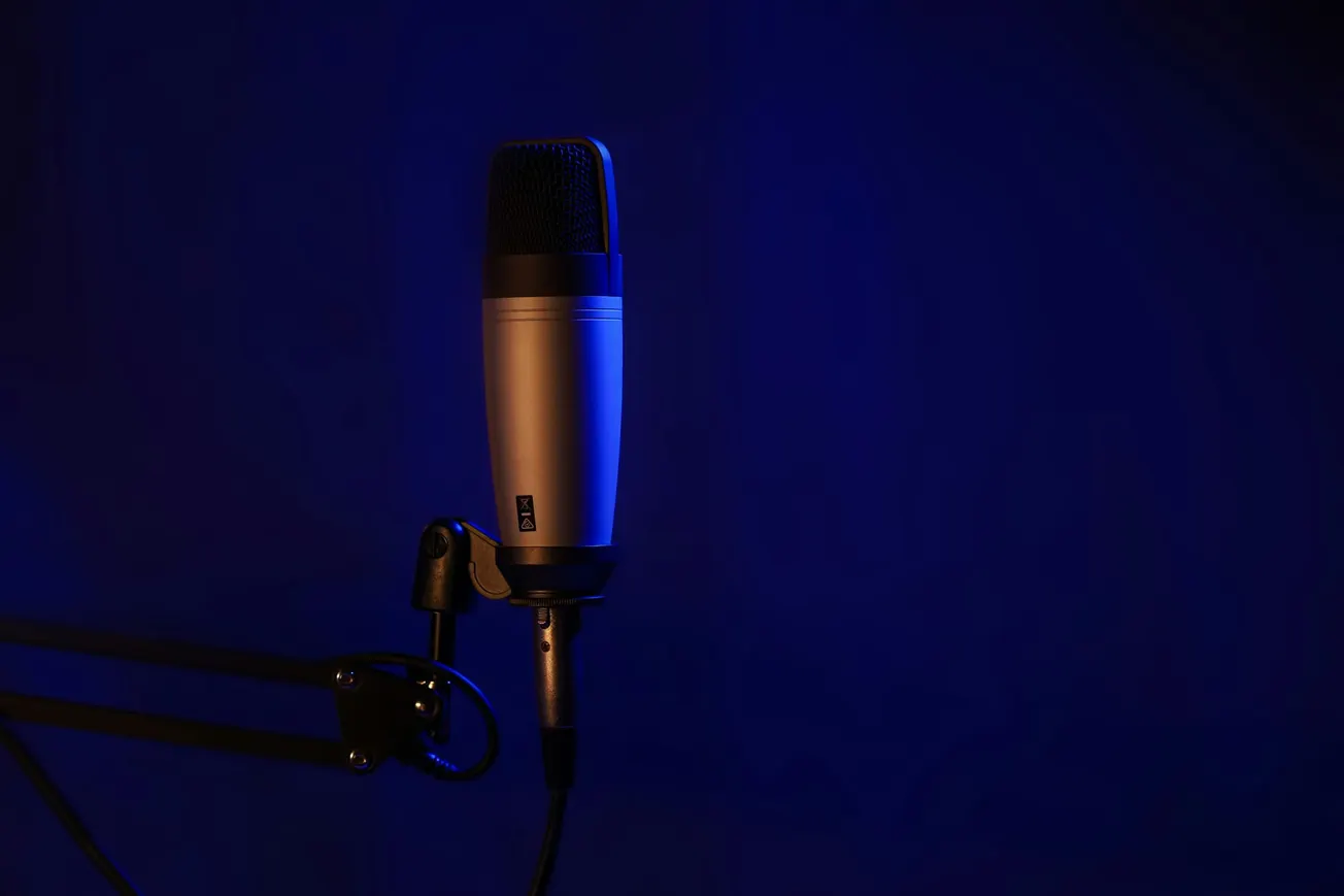 A sleek studio microphone on a stand is illuminated in soft blue light against a dark background, conveying a professional and focused atmosphere.