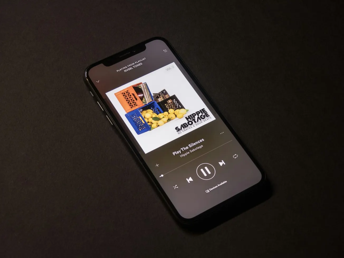 Smartphone on a dark surface displaying a music streaming app. The album cover shows crates filled with lemons. Minimalist and relaxed tone.