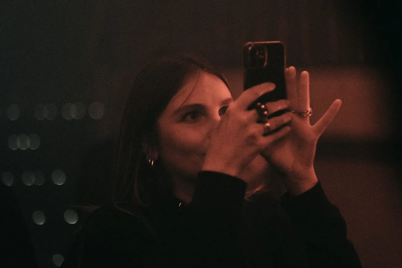 A woman in dim lighting holds a phone up, capturing a moment. The image has a warm, nostalgic grainy effect, emphasizing focus and concentration.