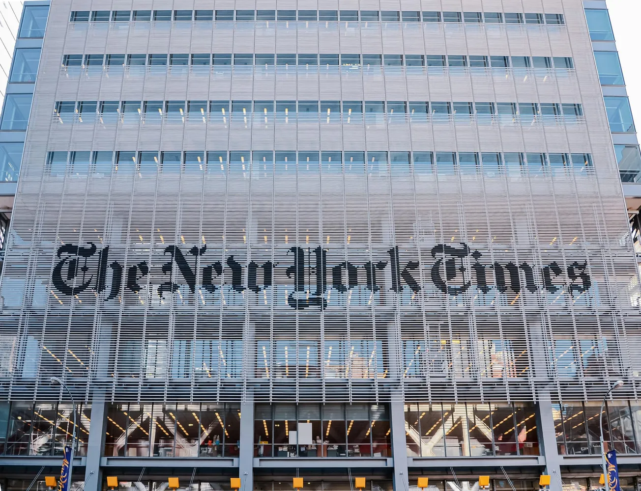 The image shows the front of The New York Times building, featuring a modern facade with large windows and the newspaper’s name in prominent black letters.