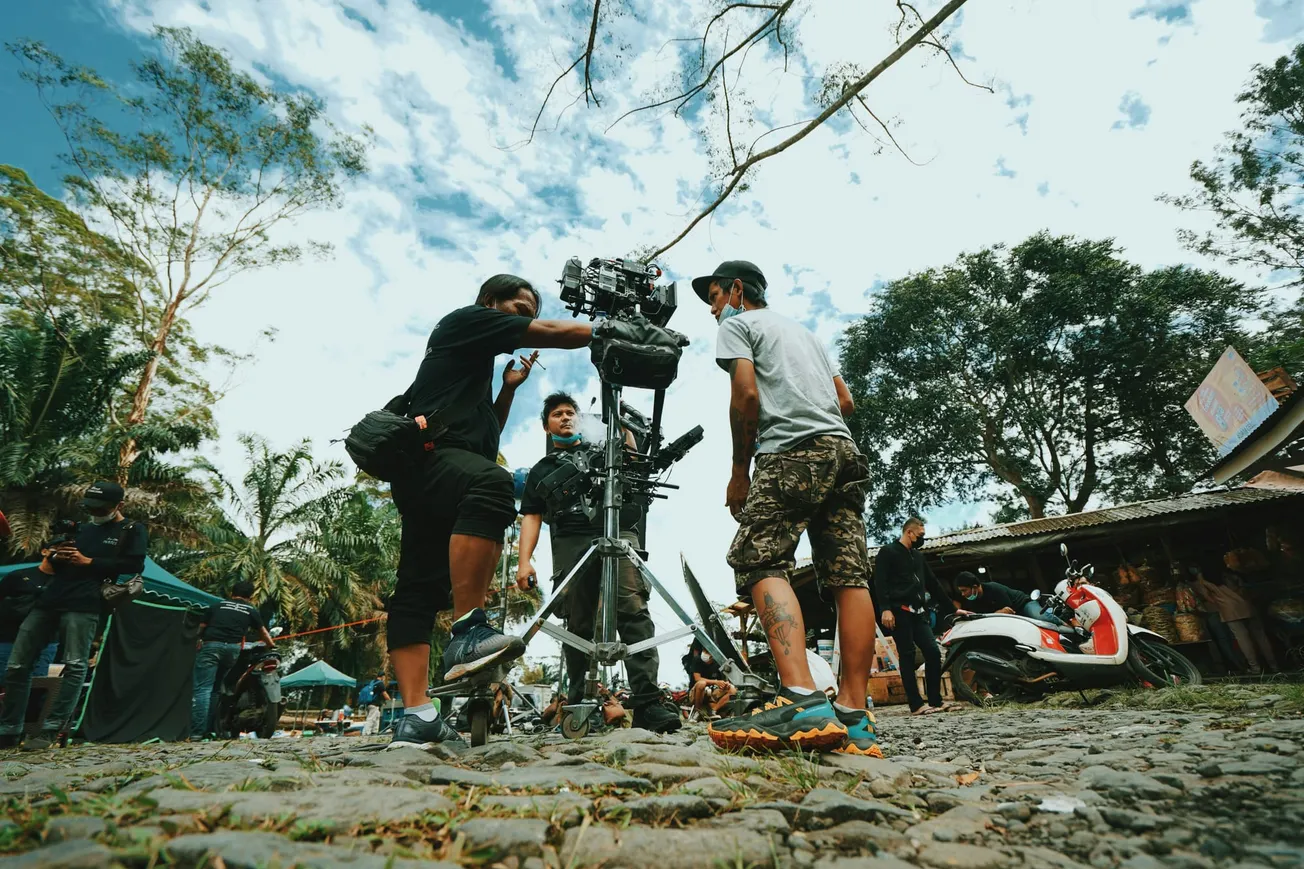 Three people operate a camera on a film set outdoors, surrounded by trees and a blue sky. A scooter is parked nearby, creating a lively, collaborative atmosphere.