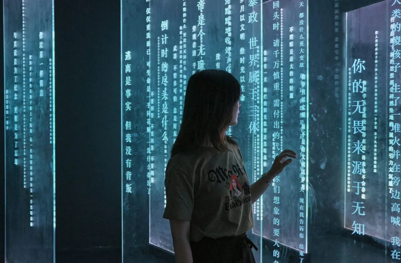A woman in a dark room examines glowing, vertical panels displaying Chinese text in soft blue light. The scene feels serene and contemplative.