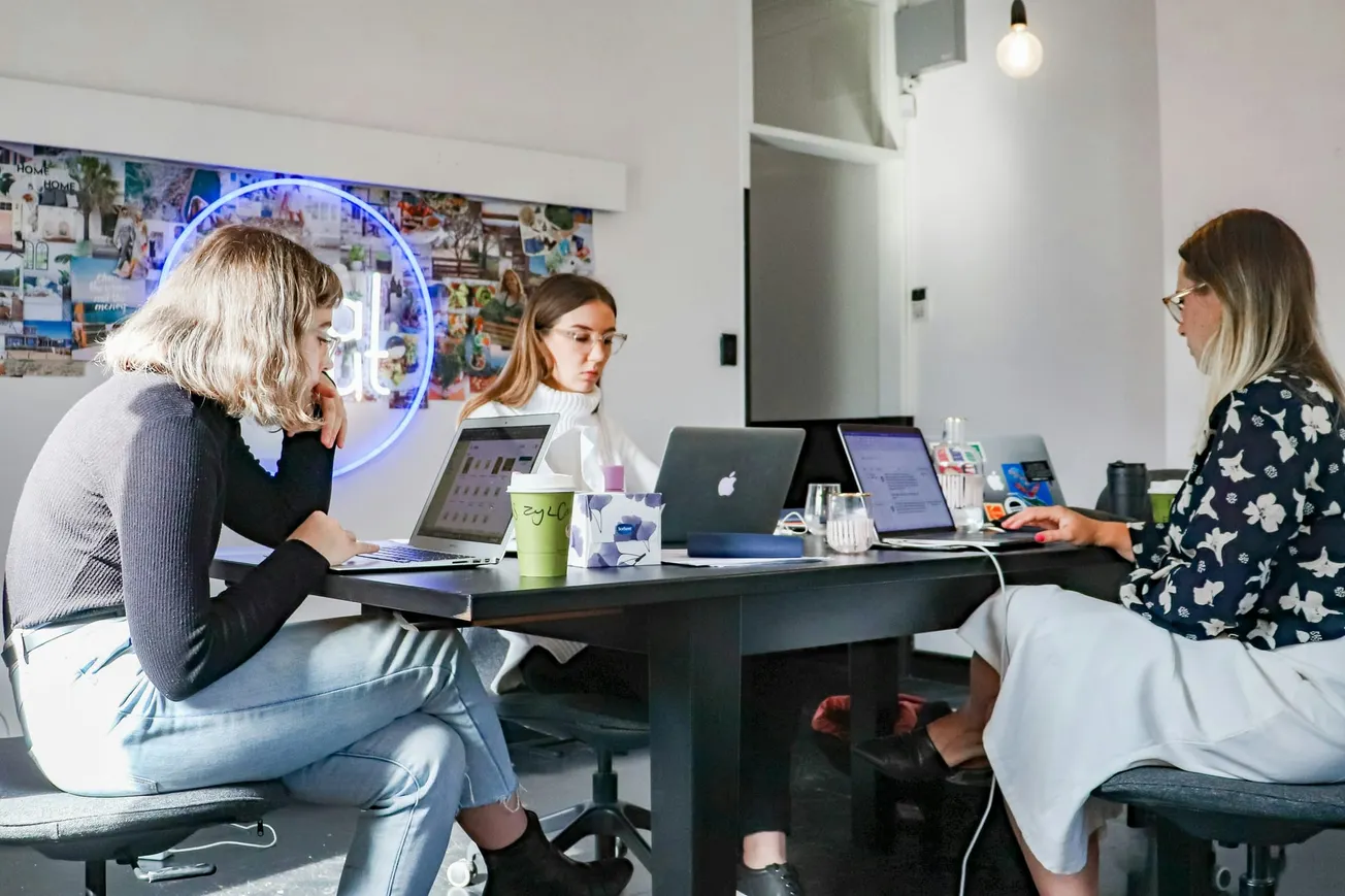 Three women are working intently on laptops at a table in a modern office. A neon sign and collage decorate the wall, adding a creative vibe.