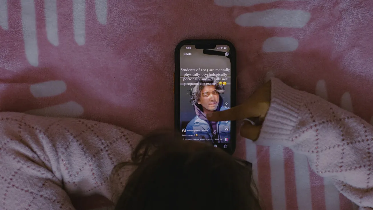 A person in a white sweater is lying on a pink blanket, looking at a smartphone. The screen shows a video with text overlay. The mood is relaxed and focused.