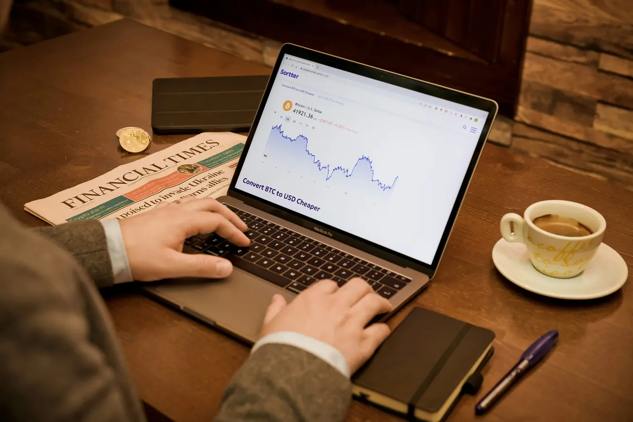 A person in a suit works on a laptop displaying a cryptocurrency chart. Nearby are a Financial Times newspaper, a notebook, a pen, and a cup of coffee on a wooden table.