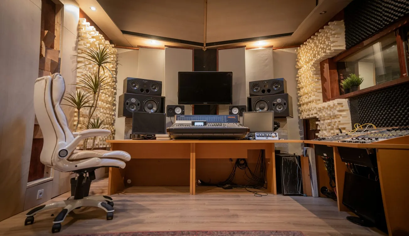 A cozy recording studio with a central mixing desk, multiple speakers, a large screen, a white swivel chair, acoustic panels, and potted plants. Warm lighting.