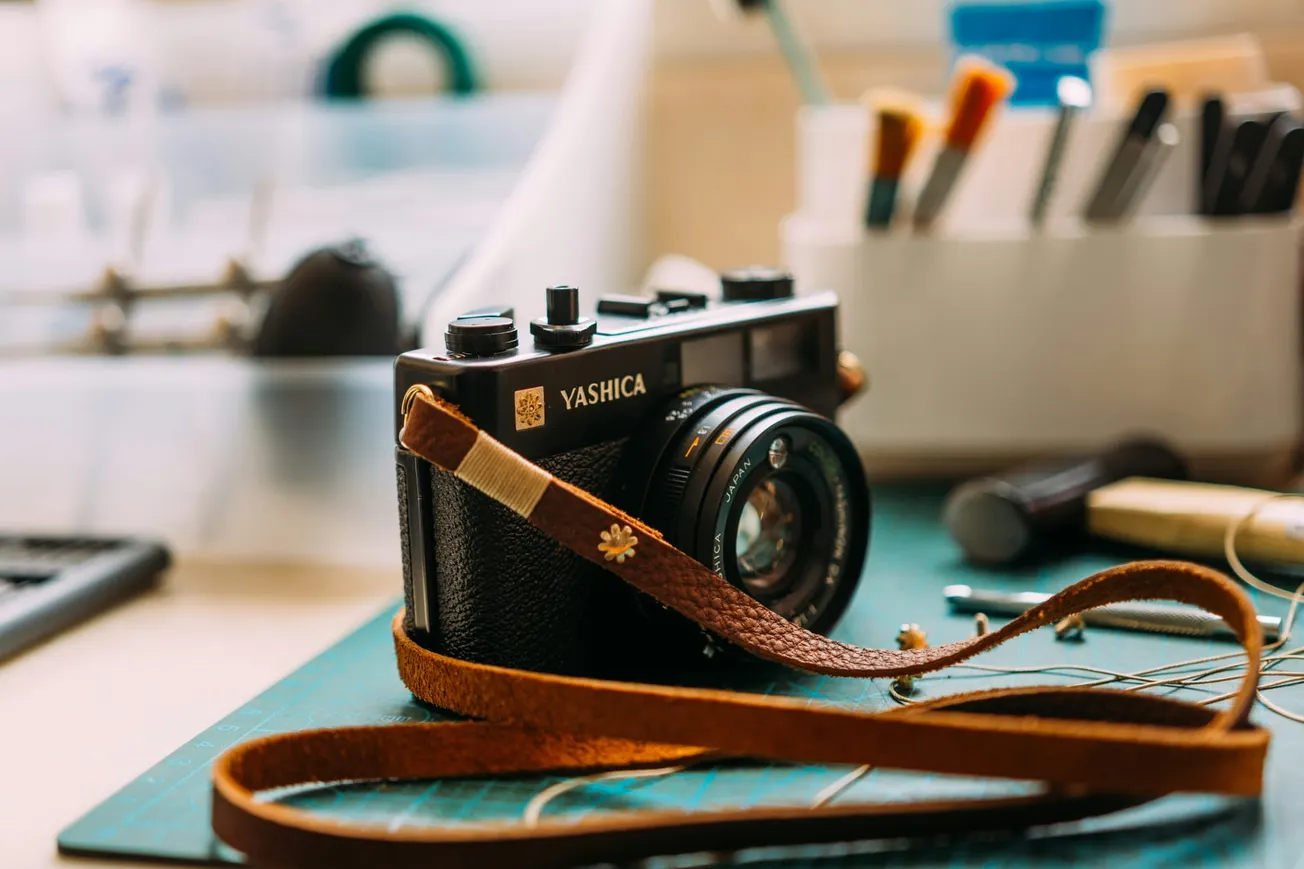 Vintage Yashica camera with a brown leather strap rests on a workspace mat, surrounded by blurred art tools, evoking a creative and nostalgic feel.