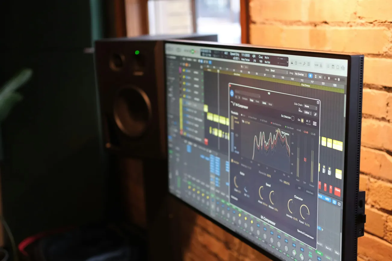 A digital audio workstation screen displays sound editing software with waveforms and controls. There's a speaker nearby. The setting is a brick-walled studio.